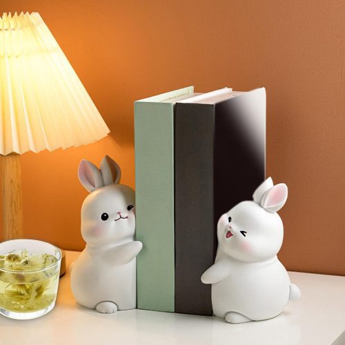 Le Serre livre lapin blanc - deux charmants serre-livres lapin en céramique - accueille des livres sur un bureau avec une lampe et des fleurs séchées, sur un mur brun clair. Un charmant accessoire de lecture pour tout amateur de livres.