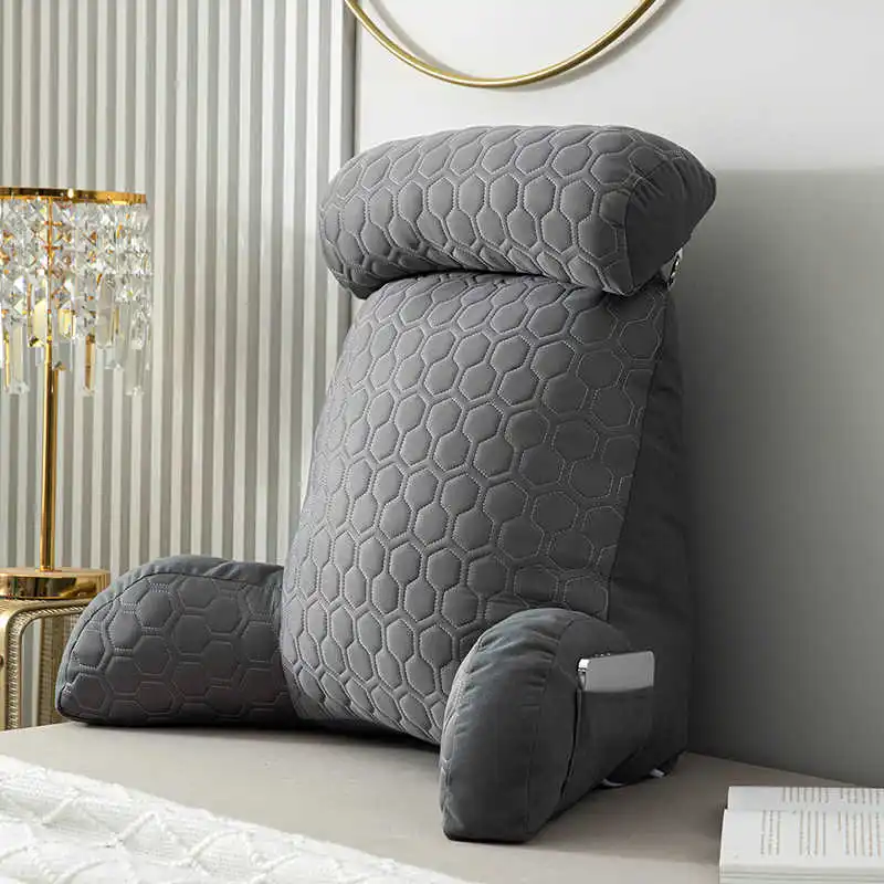 coussin de lecture book seat un grand coussin gris à motif hexagonal pour le dossier, avec des accoudoirs et un appui-tête, est placé contre un mur blanc près d'un lit - parfait pour tout passionné de lecture. une lampe en cristal et un miroir rond décoratif sont partiellement visibles à proximité.
