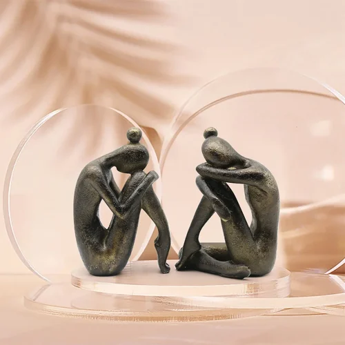 Deux sculptures abstraites en bronze représentant des personnages assis, la tête sur les genoux, reposent sur un socle rond transparent sur fond beige - idéal comme serre livre Yoda pour tout passionné de lecture ou amateur de roman.