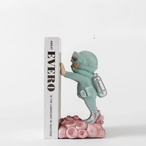 Une petite figurine "serre livre plongeur bleu" en costume sarcelle est posée sur un rocher rose, adossée à deux livres intitulés SWEAT et EVERO IS THE LUBRICANT OF SUCCESS, posés sur une étagère sur fond blanc uni.