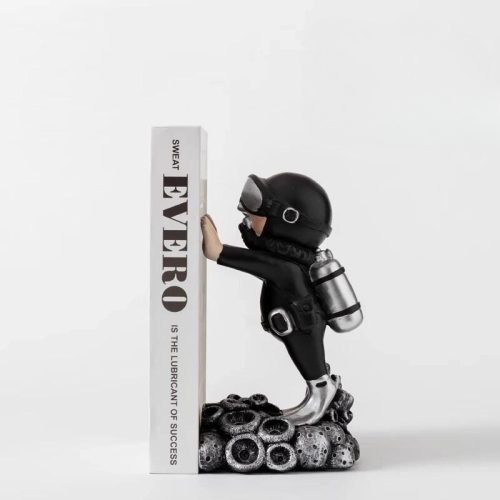 Le "serre livre plongeur argenté" représente une figurine de plongeur sous-marin en tenue noire, debout sur un corail, s'appuyant sur un serre livre vertical portant l'inscription "SWEAT IS THE LUBRICANT OF SUCCESS" - un accessoire de lecture parfait pour n'importe quelle étagère.