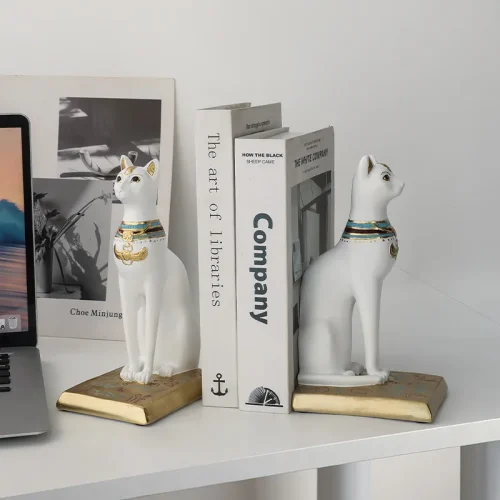 Deux serre-livres "serre livre chat égyptien" en céramique blanche avec des colliers dorés et colorés soutiennent des livres sur un bureau blanc, ajoutant une élégante touche littéraire. Un ordinateur portable et une œuvre d'art encadrée sont posés à côté des serre-livres.