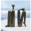 deux sculptures abstraites en bronze représentant des figures humaines assises, aux membres allongés et aux têtes rondes et sans traits, semblables à des serre livre yoda penseur, reposent sur une surface blanche, sur fond de mer bleue calme et de ciel nuageux.