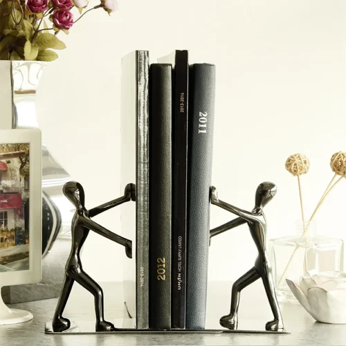 Deux serre-livres bd argentés en métal, en forme de personnages stylisés, maintiennent trois livres debout sur une surface claire. Cet accessoire élégant est idéal pour sublimer votre bibliothèque et ravir tout passionné de lecture.