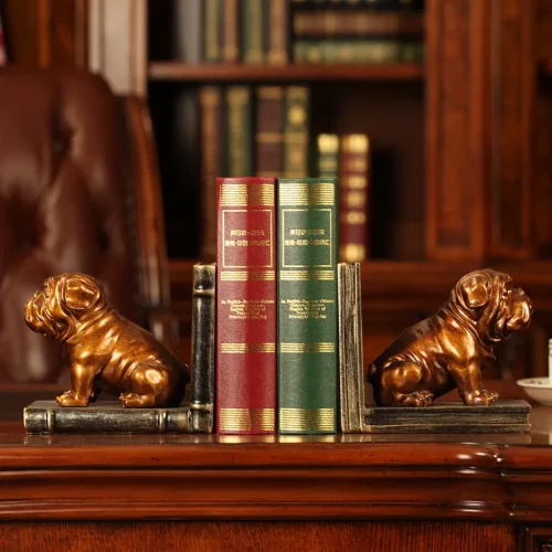 Deux serre livre chien original en bronze maintiennent trois livres-un vert et deux rouges-sur un bureau en bois, avec une bibliothèque garnie de volumes et un fauteuil en cuir marron à l'arrière, idéal pour les amoureux de lecture.