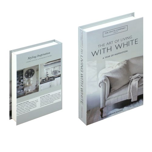 Deux livres à couverture rigide intitulés The Art of Living with White de Chrissie Rucker sont présentés sur une étagère minimaliste. L'un présente un intérieur et un canapé de couleur blanche, tandis que l'autre montre l'arrière avec une cuisine et un résumé de livre. Idéal pour les passionnés de lecture.